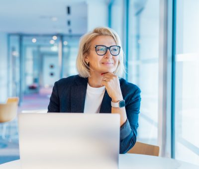 50's confident mature businesswoman dreamy looking at window, middle-aged experienced senior female professional working on laptop in open space office. Female entrepreneur working remotely